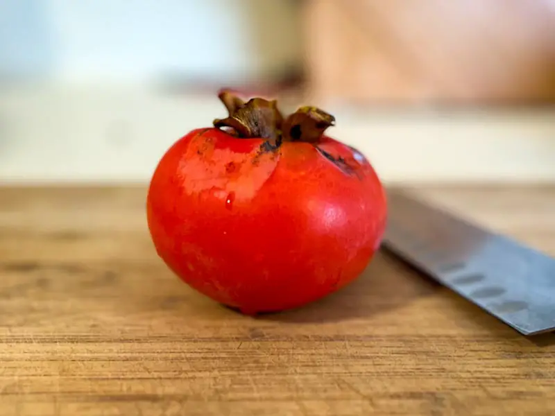 Image of ripe red-orange Fuyu persimmon on a cutting board with a knife.
