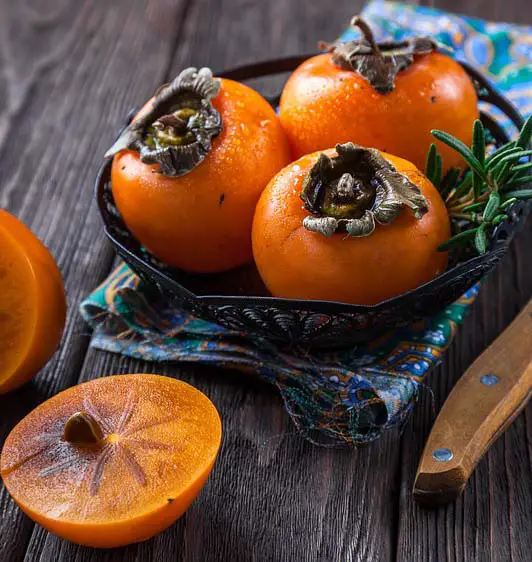 Image of ripe persimmons in a bowl with a cut persimmon to the side. The cut persimmon has dark brownish-orange flesh and a visible seed.